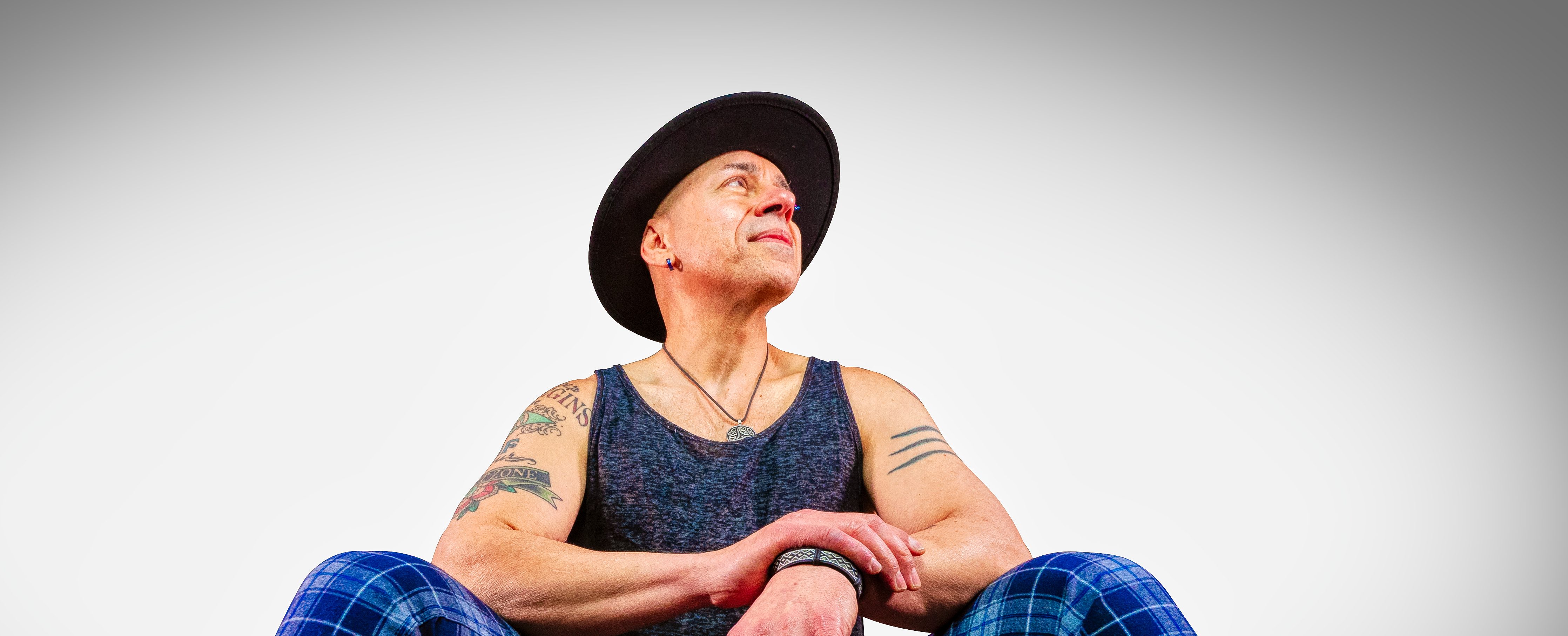 Dino Jag sits on a red seat. He wears blue and black plaid pants, a black singlet, a black wide-brimmed hat and has tattoos on his arms.