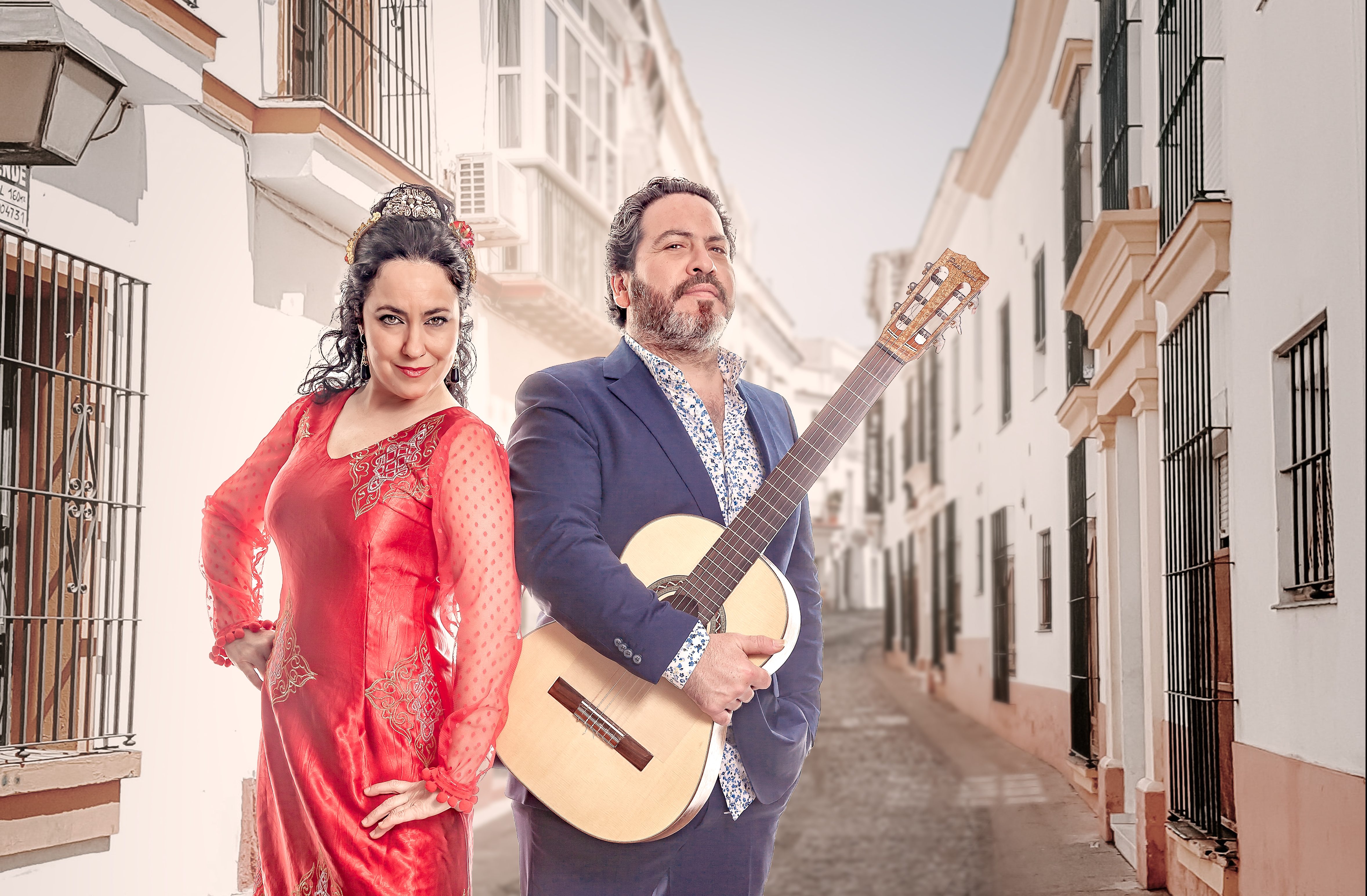 Paco and Deya Lara stand next to each other in a bright alleyway. Paco holds his guitar and Deya wears a red flamenco dress