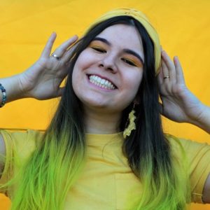 Jannah stands in front of a yellow wall. She has wears a yellow t-shirt, yellow headband and yellow earrings. The ends of her long hair are also died yellow.