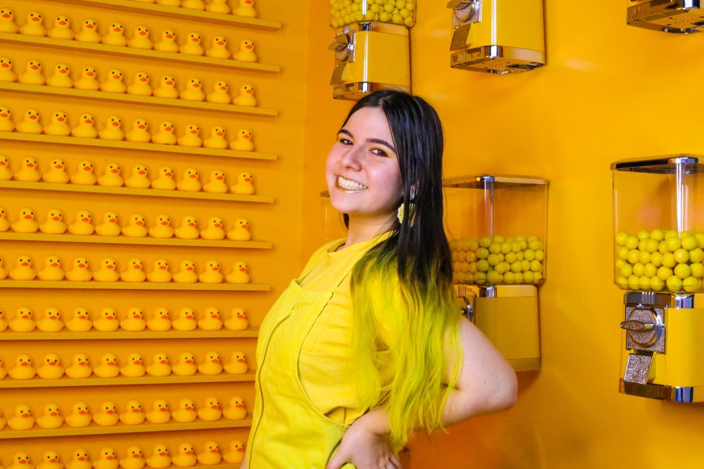 Jannah Fahiz stands in front of a yellow wall with gum ball machines. On another wall are yellow rubber ducks.