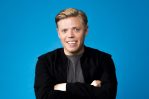 Rob Beckett looks at the camera. He is smiling and wears a grey t-shirt and black jacket. The wall behind him is light blue.