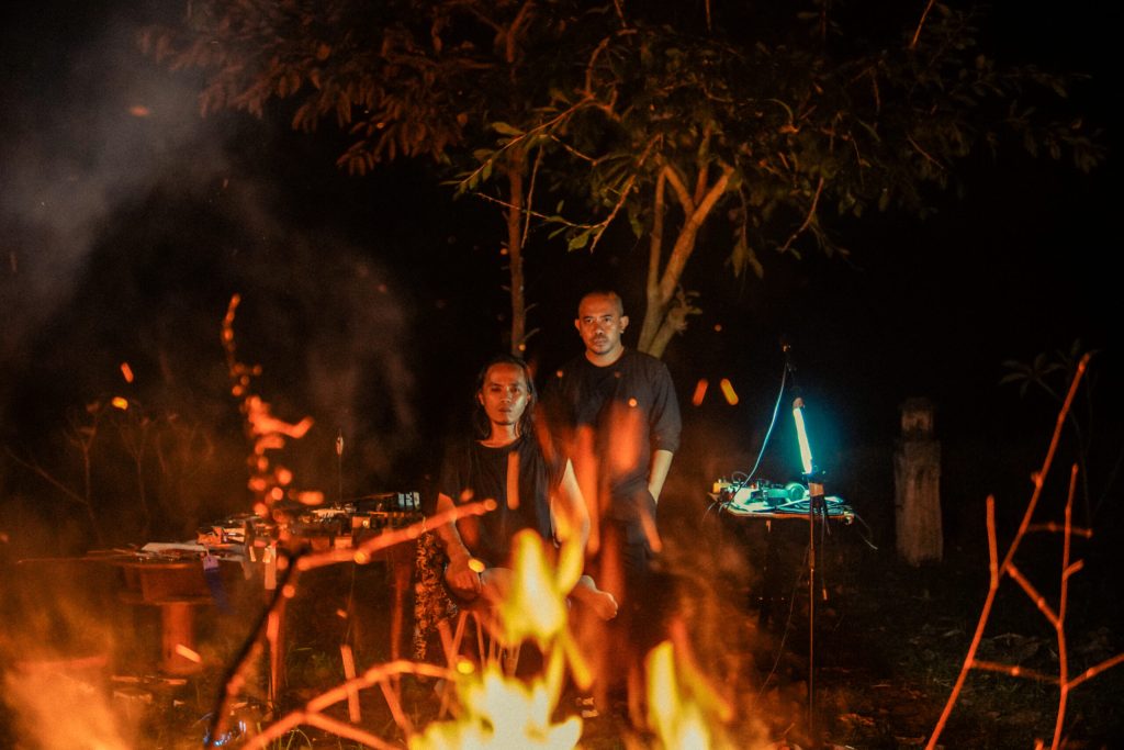 Two people staring into a fire. There is electronic music equipment around them.