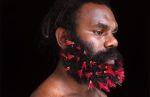 Man with black beard is photographed looking to his left, his body is also facing left. He has red feathers weaved through hi beard.