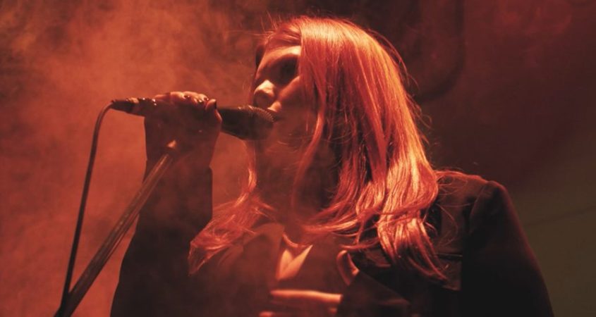 Bayley Pearl sings into a microphone. There photo is tinted red and there is staged smoke in the background. Bayley has long hair and wears a long-sleeved black top/