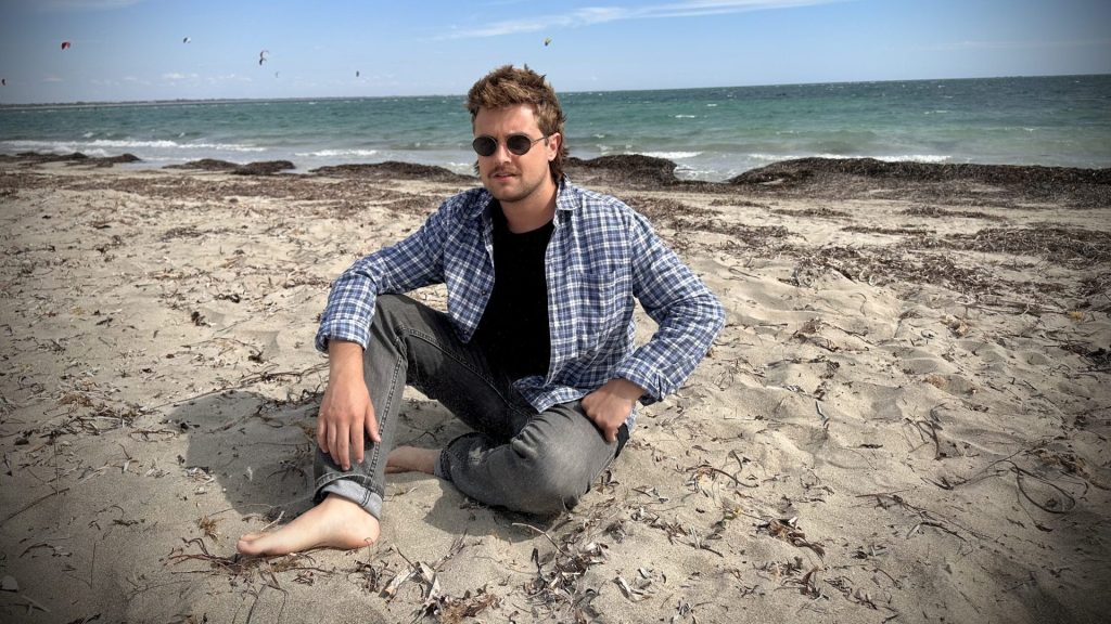 Man sitting on the beach with the ocean and blue sky behind him. He is facing the camera wearing sunglasses, jeans, blue checked shirt and no shoes.