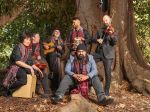 The ensemble Farhan Shah & Sufi-Oz are sitting at the base of a large tree. They are holding their instruments and smiling