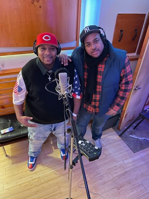 Two men, the one on the left, Asim I.I. They appear to be in a studio and are wearing baseball caps and headphones. they are smiling at the camera.