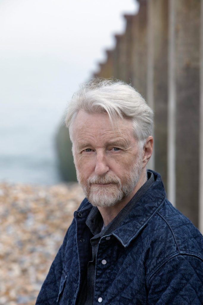 Billy Bragg is sitting and looking at the camera with intensity with his blue eyes. He is wearing in a blue jacket and shirt.
