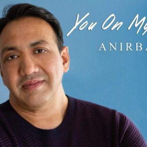 A photo of Anirban Jee's single cover. He is leaning against a blue wall and smiling