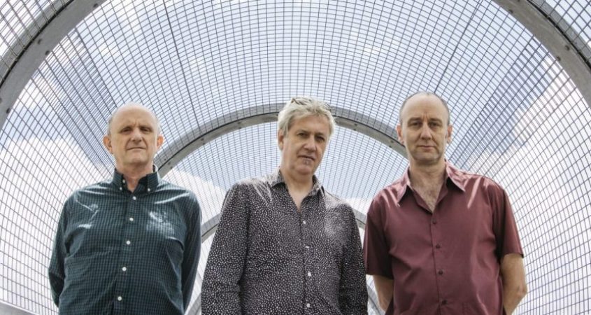 The three members of The Necks are standing outside on a covered walkway, looking at the camera. They are wearing collard shirts.
