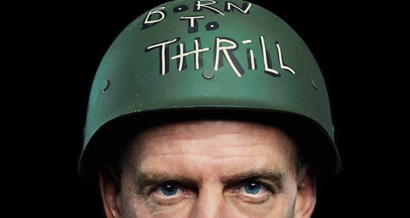 A portrait of Fatboy Slim looking at the camera with his blue eyes and wearing a green helmet that says 'Born to Thrill'