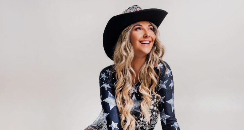A portrait of Kayla Krystin against a white studio backdrop. She is wearing a fun country music style outfit with a star print. She is smiling and her long blonde hair frames her face.