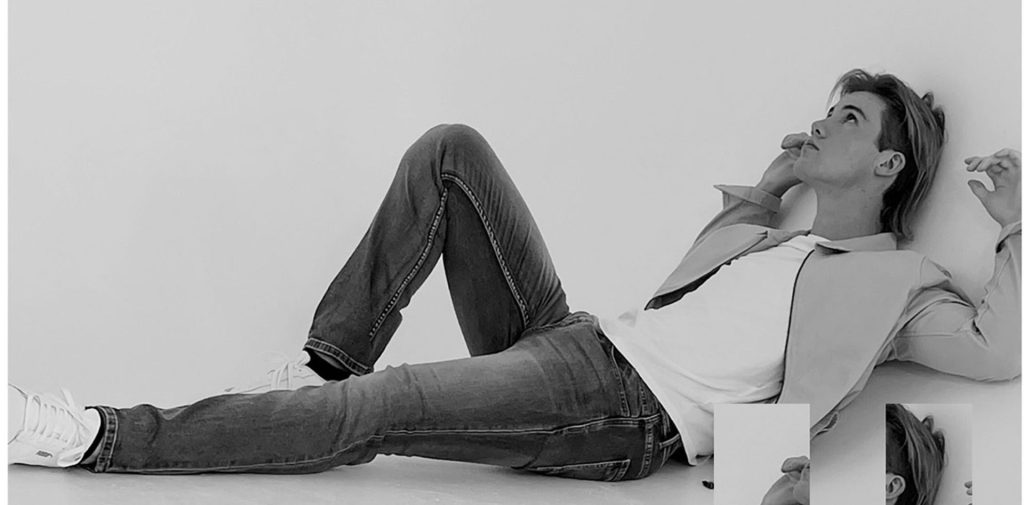 A black and white studio portrait of Jaxon Reid. He is laying down, looking up and wearing jeans and a shirt.