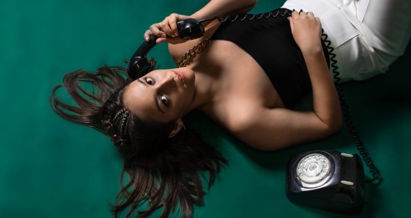 A portrait of Rachael Coltrona. She is lying down on a green floor and talking on a black rotary phone. She is wearing a black halter top and white pants with her hair flowing on the floor.